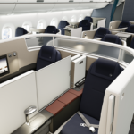Se aprecian varios asientos de avión, separados en cubículos, en una cabina de primera clase en cuya pared trasera se aprecia el logo de Lufthansa.