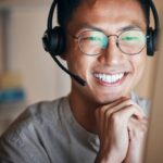 Joven con anteojos mira la pantalla de un computador mientras sonríe. Usa una diadema con audífonos y un micrófono incorporado.