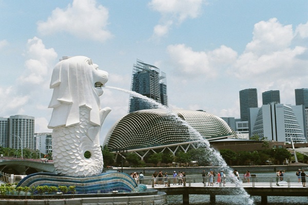 Una figura blanca de piedra, con cabeza de león, y torso y cola de sirena, vierte agua en una fuente. Al fondo, se ven diversos edificios de la ciudad-estado de Singapur.