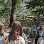 Un grupo de scouts, conformado por seis niños y un adulto, recorren a pie una zona boscosa.