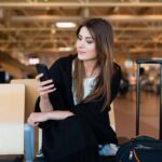 Mujer joven sonríe mientras mira la pantalla de su celular en la sala de espera de un aeropuerto.