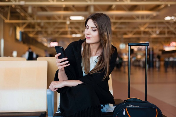 Mujer joven sonríe mientras mira la pantalla de su celular en la sala de espera de un aeropuerto.