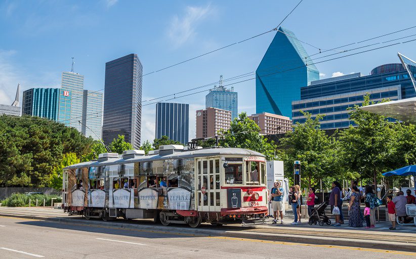 Un trolebús se acerca a una estación del McKinney Avenue Trolley, en Dallas, Texas. Al fondo, se ven varios edificios de la ciudad.